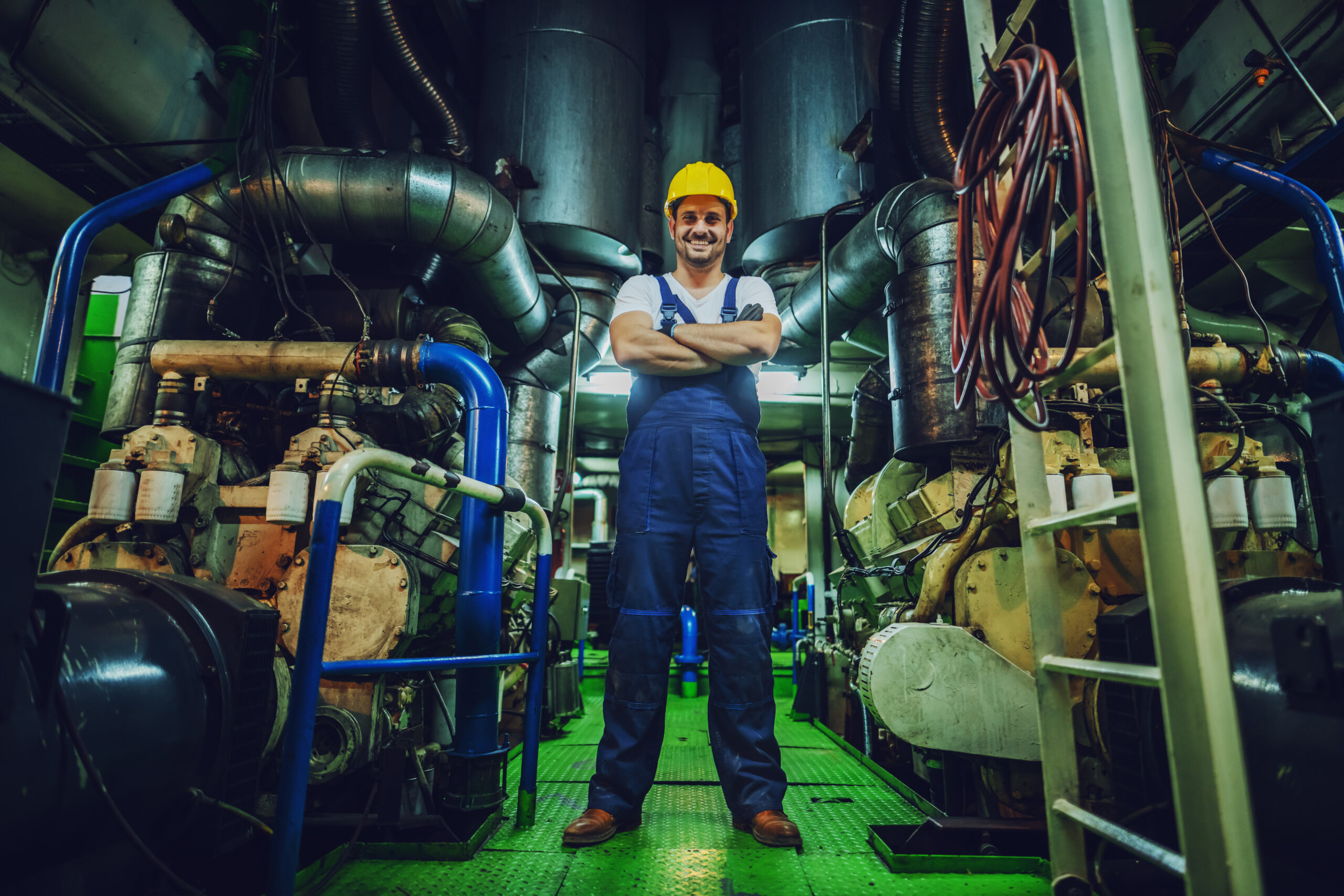 Low angle view of handsome caucasian worker in overall and helmet on head standing in ship next to engine with arms crossed.